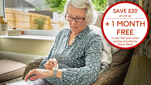 An older woman with white hair and glasses sits in a wicker armchair by a window, looking down at her digital personal alarm
