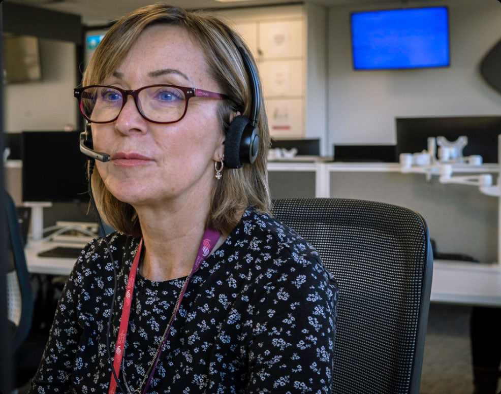 A woman with short blonde hair, glasses, and a headset sits in an office environment wearing a black floral-patterned top and a pink lanyard.