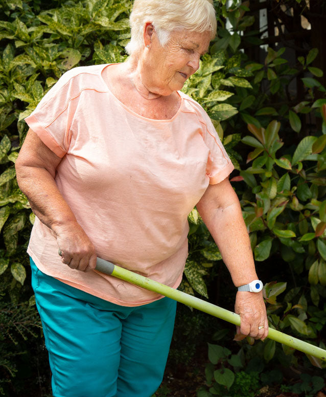 An older woman in a garden wearing a fall alarm and holding a long green garden tool handle. 