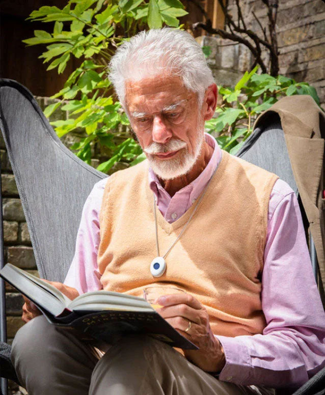 An older man with white hair and a beard sits in a garden chair, reading a book. He is wearing a fall alarm