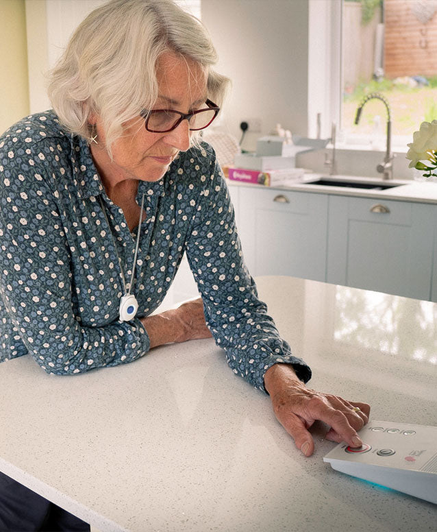 An older woman with white hair and glasses is leaning over a kitchen island, pressing buttons on an alarm base unit