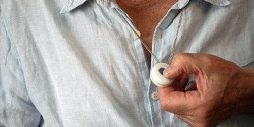 A close-up of a person wearing a light blue button-up shirt and holding a small round white pendant necklace with a blue circular detail.